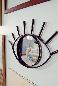 an eye shaped mirror hanging on the side of a white wall next to a painting