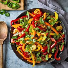 a salad with peppers and green onions in a black bowl next to a wooden spoon