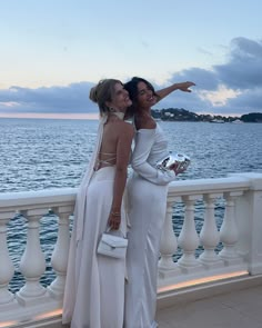 two women standing next to each other on a balcony near the ocean and one is pointing at something