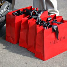 three red shopping bags with black ribbons tied to them, sitting on the ground in front of a woman's feet