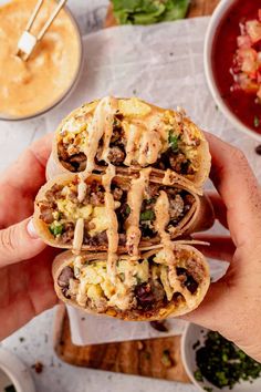 a hand holding a burrito filled with meat, cheese and other toppings next to bowls of soup