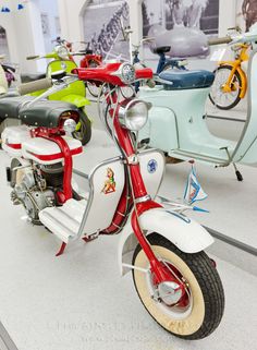 several scooters are lined up on display in a museum room with other scooters behind them