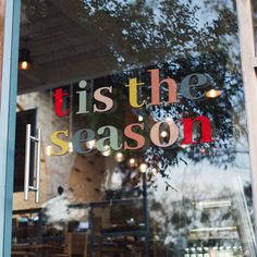 the window of a restaurant that says tis the season in multicolored letters on it