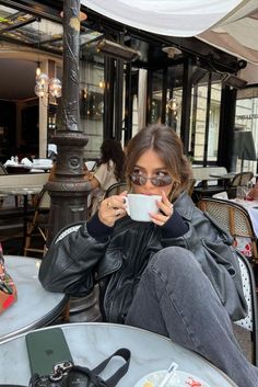 a woman sitting at an outdoor table drinking from a coffee cup and looking into the camera