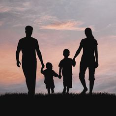 the silhouette of two adults and two children holding hands as they walk across a field at sunset