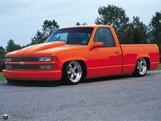 an orange pick up truck parked in a parking lot next to some grass and trees