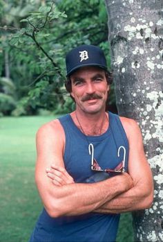 a man wearing a hat and glasses standing next to a tree with his arms crossed