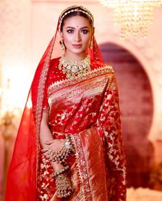 a woman in a red and gold bridal outfit with jewelry on her head, standing next to a chandelier