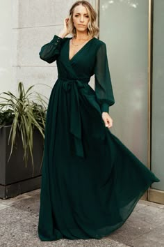 a woman in a long green dress standing on the sidewalk with her hands behind her head