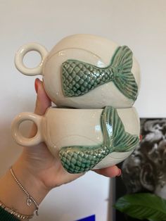 two ceramic mugs with fish on them are being held by a woman's hand