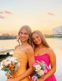 two women in dresses standing next to each other near the water with their arms around each other