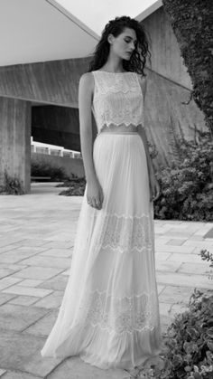 a woman in a long white dress standing next to a tree and bushes with her hand on her hip