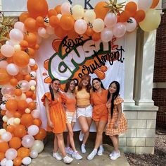 four girls posing in front of an orange and white balloon arch