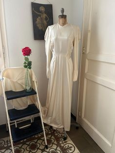 a white dress on display next to a chair and vase with a rose in it