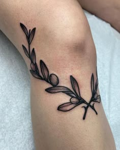 an olive branch tattoo on the leg is shown in black and grey ink, with leaves around it