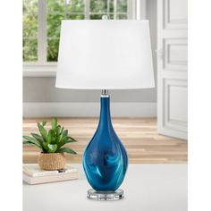 a blue glass table lamp with a white shade on the base and a potted plant next to it