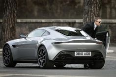 a man in a suit sitting on the back of a silver sports car with its door open