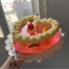 a person holding up a cake in front of a window with a cherry on top