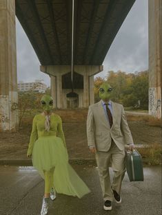 two people dressed in alien costumes walking under a bridge with suitcases on their feet