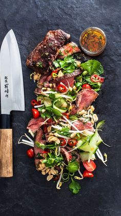 a salad with meat and veggies next to a knife on a black surface