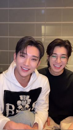 two young men sitting next to each other in front of a tiled wall with white tiles