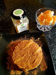 a pancake on a black plate next to some oranges and seasoning in a glass bowl