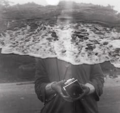 a man holding a camera standing in front of the ocean with waves crashing on him