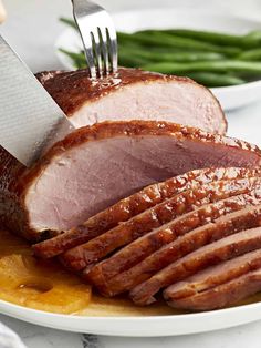 a ham being cut with a knife and fork on a plate next to green beans