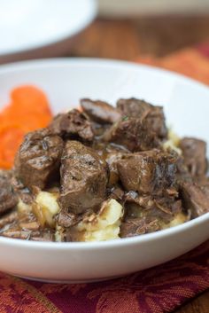 a white bowl filled with meat and potatoes on top of a table next to carrots