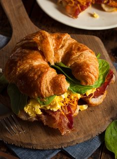 a croissant sandwich with bacon, cheese and spinach on a wooden cutting board