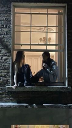 two people sitting on a window sill in front of a brick building at night