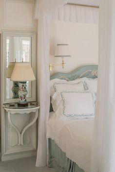 a white bed sitting under a window next to a lamp on a table with a mirror