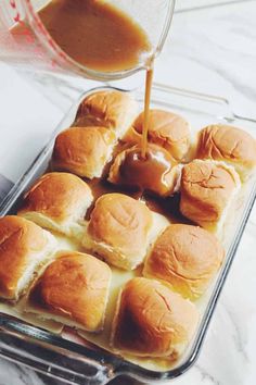 someone pouring sauce on top of rolls in a baking pan with other rolls and buns