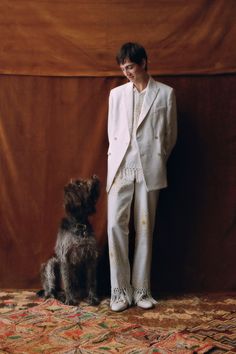 a woman standing next to a dog in front of a brown wall wearing a white suit