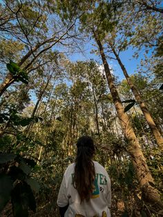 a woman standing in the middle of a forest looking up into the sky at tall trees