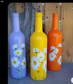 three painted bottles sitting next to each other on a cement floor near a fence and tree