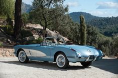 an old blue sports car parked in front of some rocks and trees with mountains in the background