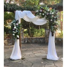 an outdoor wedding setup with white draping and flowers on the arch, surrounded by greenery