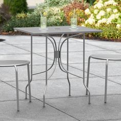 a table and two chairs sitting on top of a cement floor next to bushes with flowers in the background