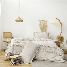 a bed with white sheets and pillows in a room next to a chair, lamp and books on the floor