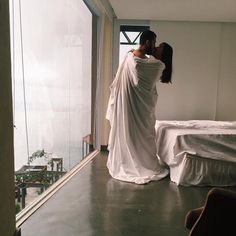 a woman in a white dress standing next to a bed and looking out the window