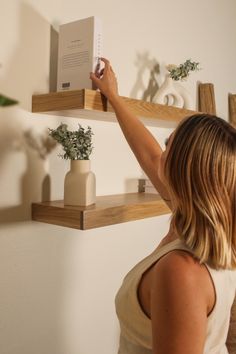 a woman is holding up a book in front of some shelves with plants on them