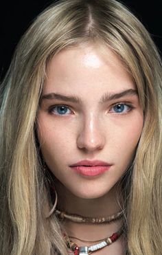 a woman with long blonde hair and blue eyes wearing necklaces on her neck is looking at the camera