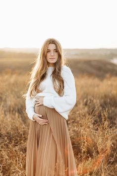 a pregnant woman standing in a field with her hands on her stomach wearing a white sweater and brown pleated skirt