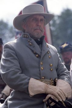 an old man in uniform sitting on a horse with other men standing behind him and looking off into the distance