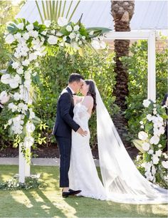 a bride and groom kissing under an arch decorated with white flowers at their wedding ceremony