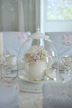 a glass tea pot with flowers in it sitting on a table next to cups and saucers