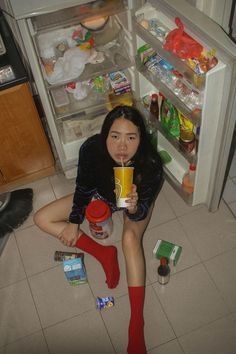 a woman sitting on the floor in front of an open refrigerator drinking from a cup