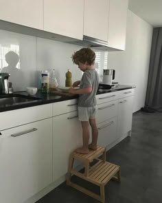 a young boy standing on a step stool in a white kitchen with black countertops