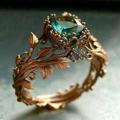 a gold ring with an emerald colored stone surrounded by leafy vines and leaves, on a gray surface